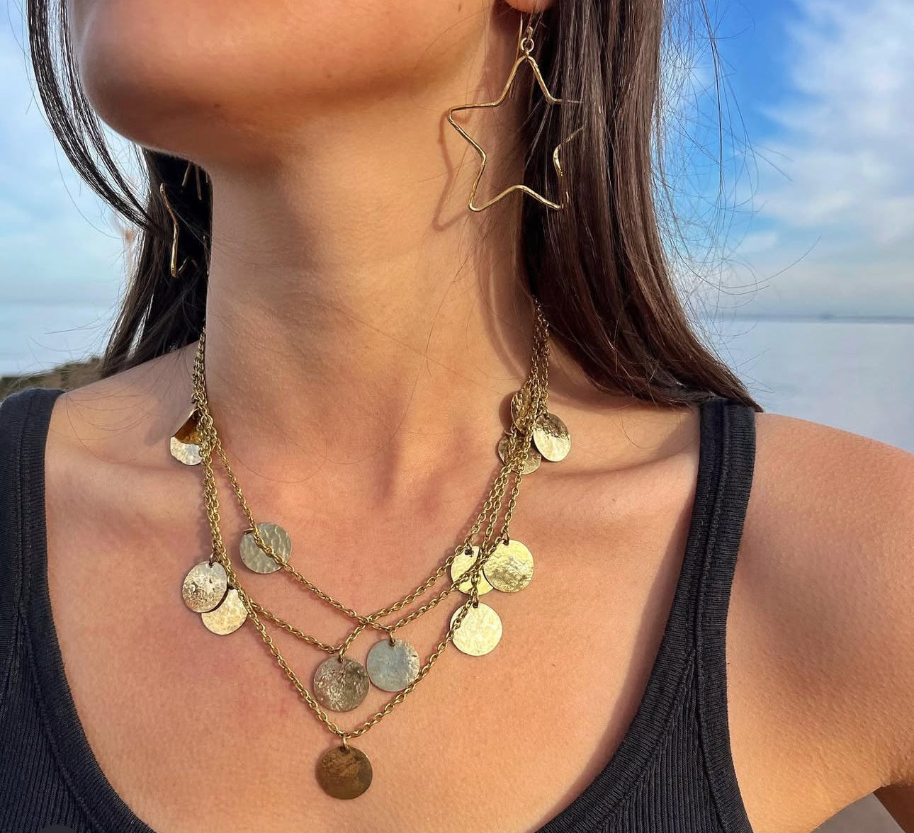 Close-up of woman wearing a layered gold coin chandelier necklace with matching star-shaped earrings against natural beach backdrop, showcasing the hammered disc texture and multi-strand design