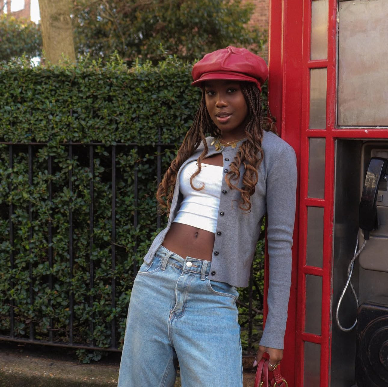 Woman with long braids wearing a delicate gold coin necklace paired with casual outfit of white crop top, gray cardigan and light denim, standing beside a classic red telephone booth